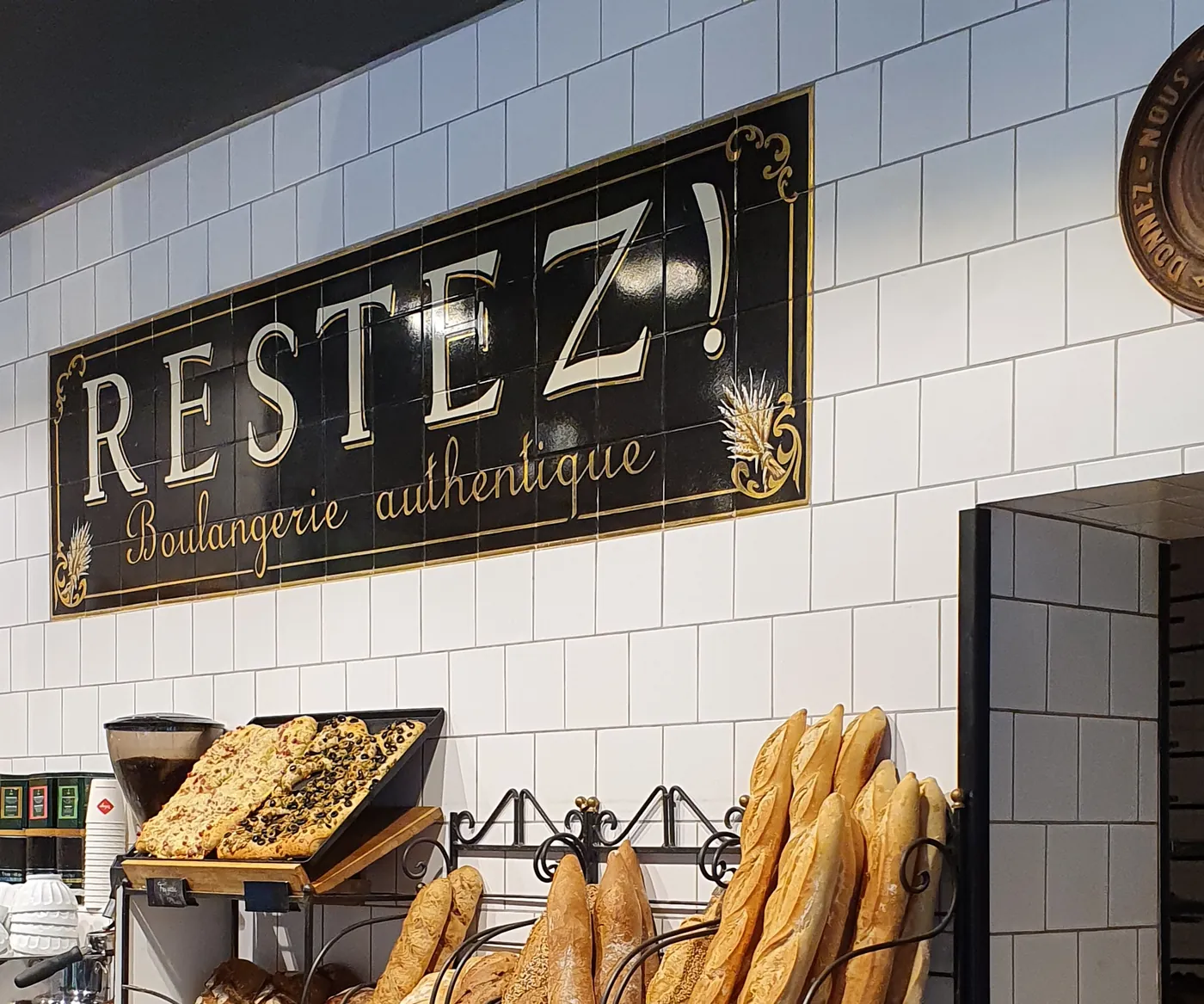 Baguettes im schmiedeeisernen Gestell vor weißen Fliesen, darüber das Schild „Restez! Boulangerie authentique“