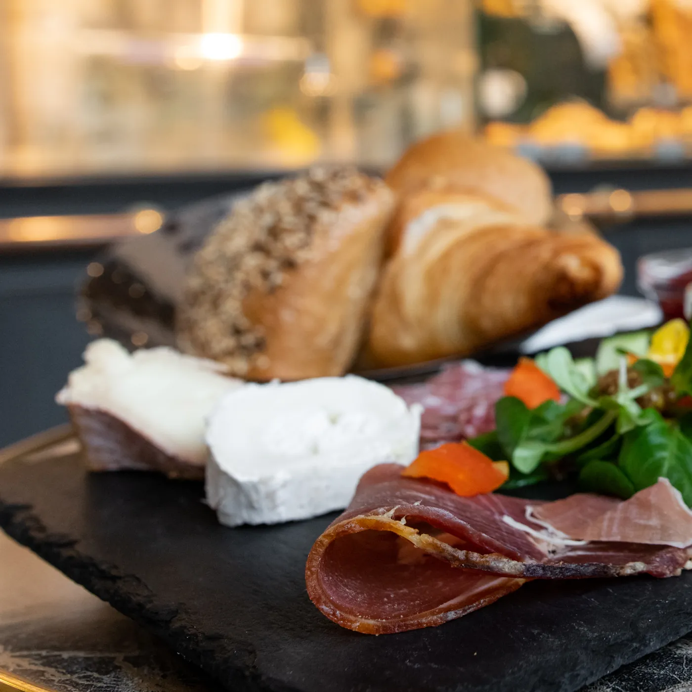 Rohschinken, Ziegenkäse und Feldsalat auf einer Schieferplatte, dahinter Körnerbrötchen und Croissant