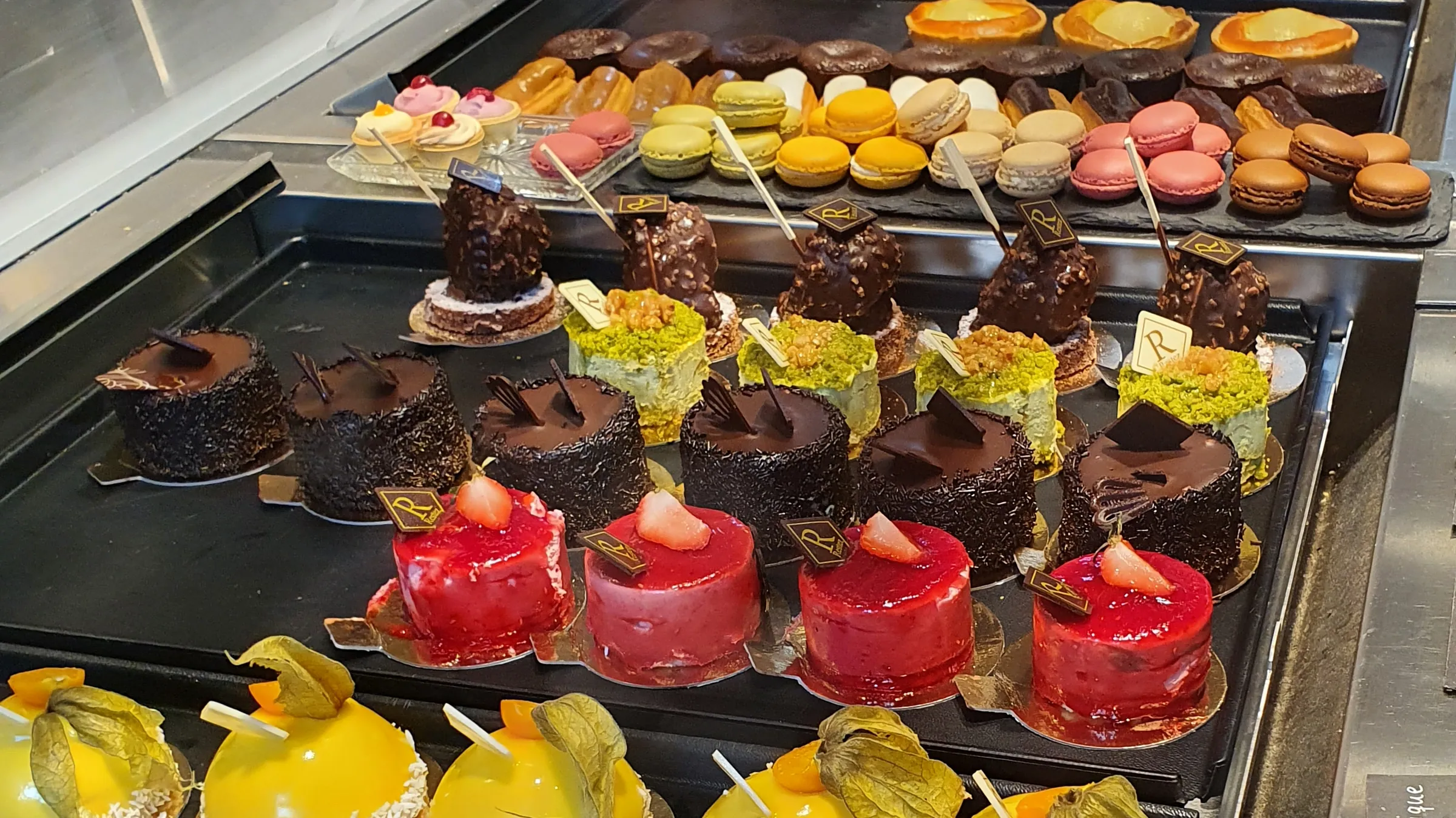 Vitrine mit Törtchen in Reihen: Himbeer, Schokolade, Pistazie und Mango, dahinter Macarons und Tartes